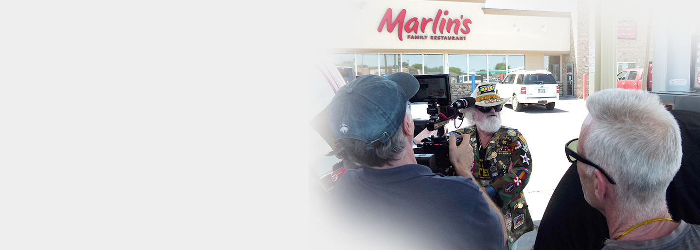 A man dressed in a colorful, patch-covered jacket and wearing a wide-brimmed hat with decorative pins is being filmed outside a restaurant named "Marlin's Family Restaurant" by a cameraman and another person. The scene is set in a parking lot with cars visible in the background.