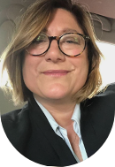 Close-up of a smiling woman with short brown hair, glasses, wearing a dark blazer and a light-colored shirt, sitting in a vehicle.