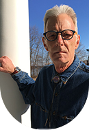An older person with gray hair, glasses, and a denim jacket standing outdoors against a blue sky and trees.