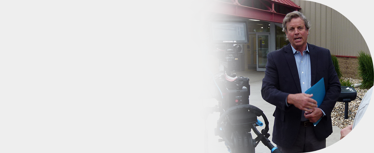 A man in a business suit holding a blue folder being interviewed outside a building with cameras recording him.