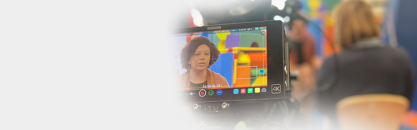 A person operating a professional video camera, filming a woman with curly hair in an orange top against a colorful abstract background.