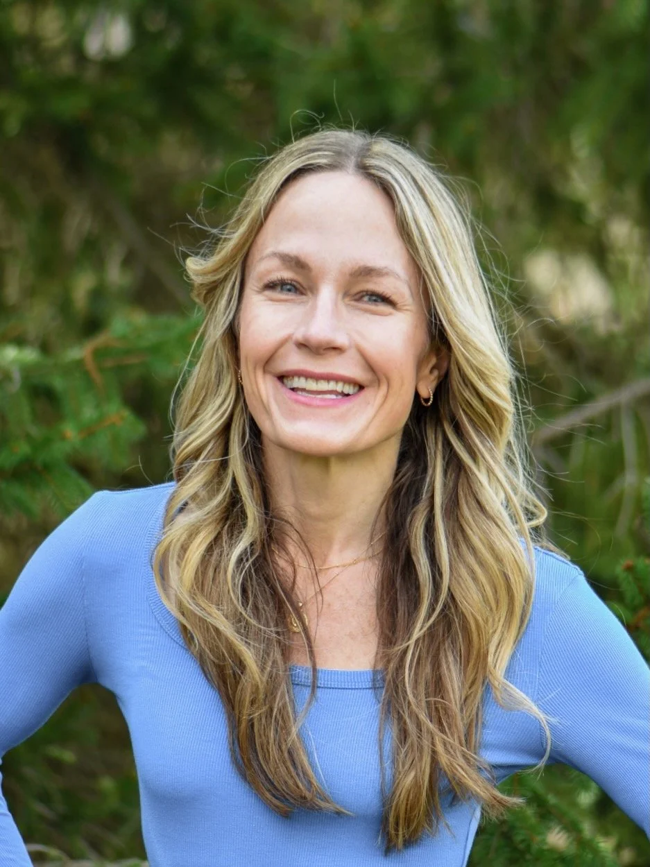 A smiling woman with long, wavy blonde hair, wearing a blue long-sleeve shirt, standing outdoors with green foliage in the background.