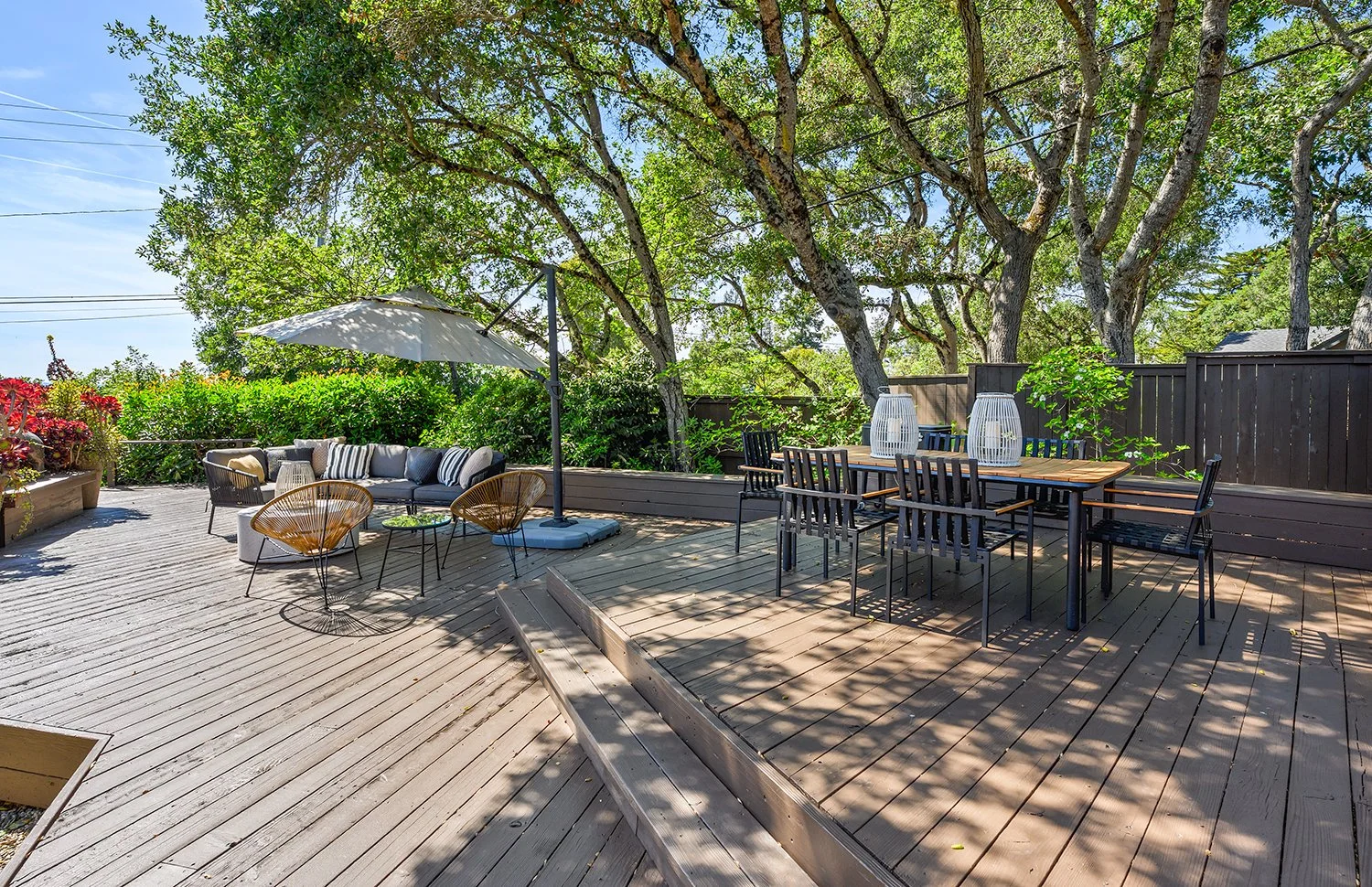 A spacious outdoor wooden patio with a seating area, an umbrella, a dining table with chairs, and trees providing shade.
