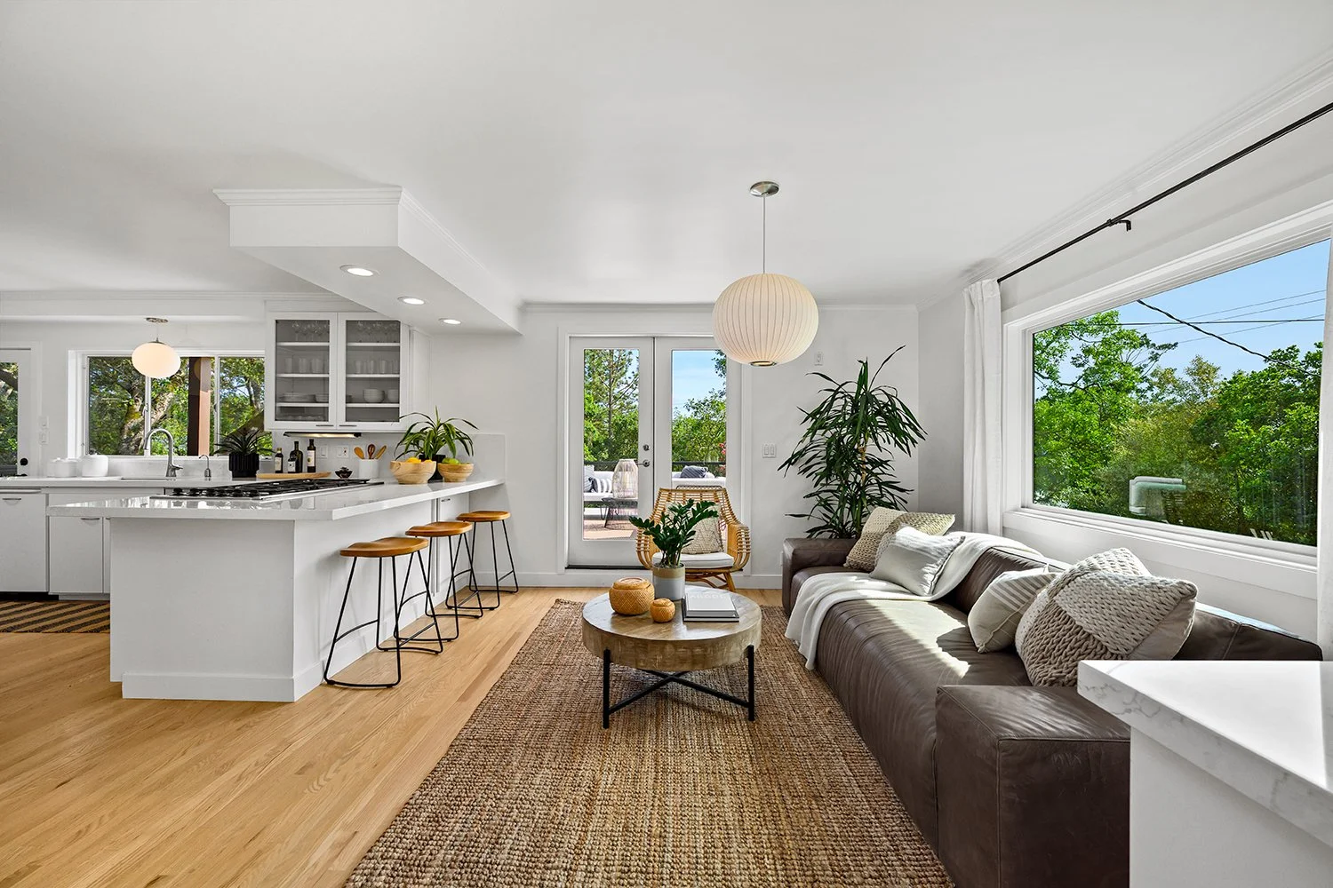 Bright living room with large window showing green trees, white walls, and wooden flooring. Light-colored sofa with throw pillows, wooden coffee table with decorative items, and indoor plants. Open kitchen with white cabinets and wooden stools.