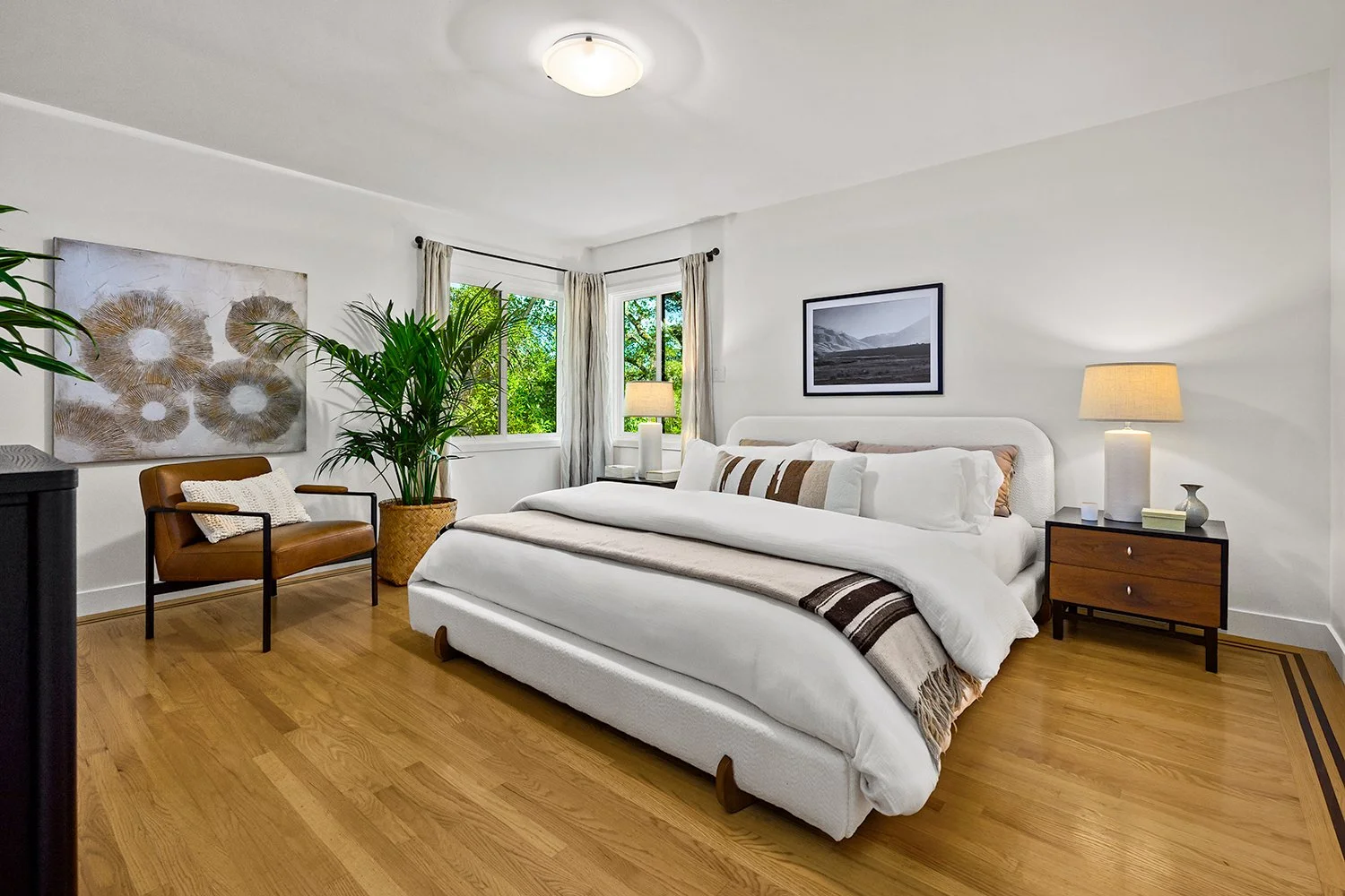 The bedroom features a white bed with neutral colored pillows, wooden nightstands with lamps, a brown leather armchair, a large potted plant, and windows with white curtains providing natural light.