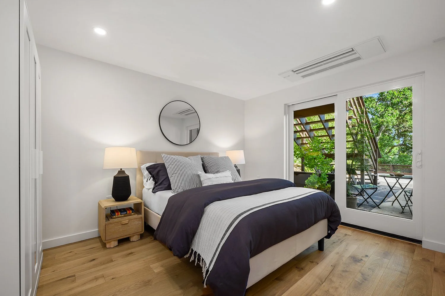 A modern bedroom with a bed, nightstand, lamps, and a large window leading to an outdoor balcony with trees.