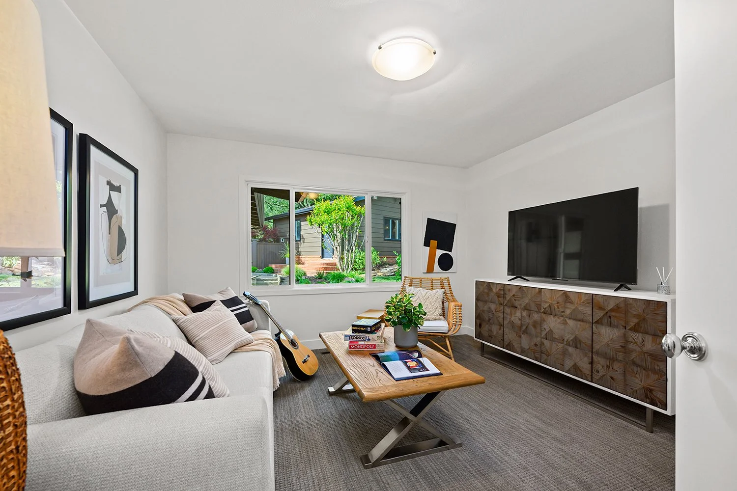 Cozy living room with white sofa, black and white pillows, wooden coffee table with books and plant, wicker chair, large window showing green outdoor yard, flat-screen TV on wooden sideboard, abstract artwork on wall, and gray carpeted floor.