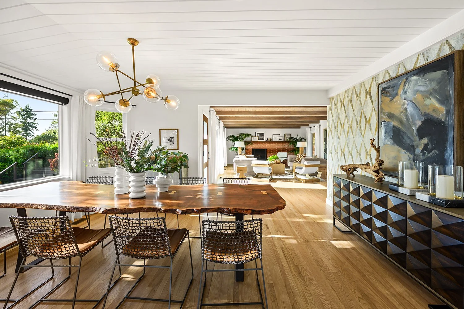 Open living and dining area with wooden floors, a wooden dining table, six metal and leather chairs, a modern chandelier, white curtains, large windows, and a living room with white furniture, a fireplace, and framed pictures.