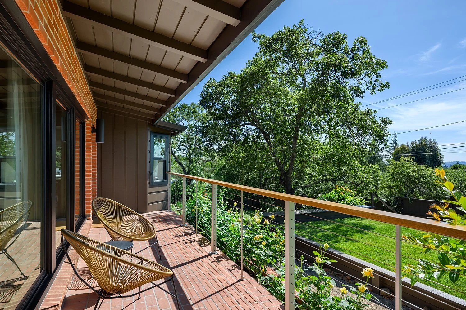 Balcony with three chairs overlooking a backyard with trees and green grass on a sunny day.
