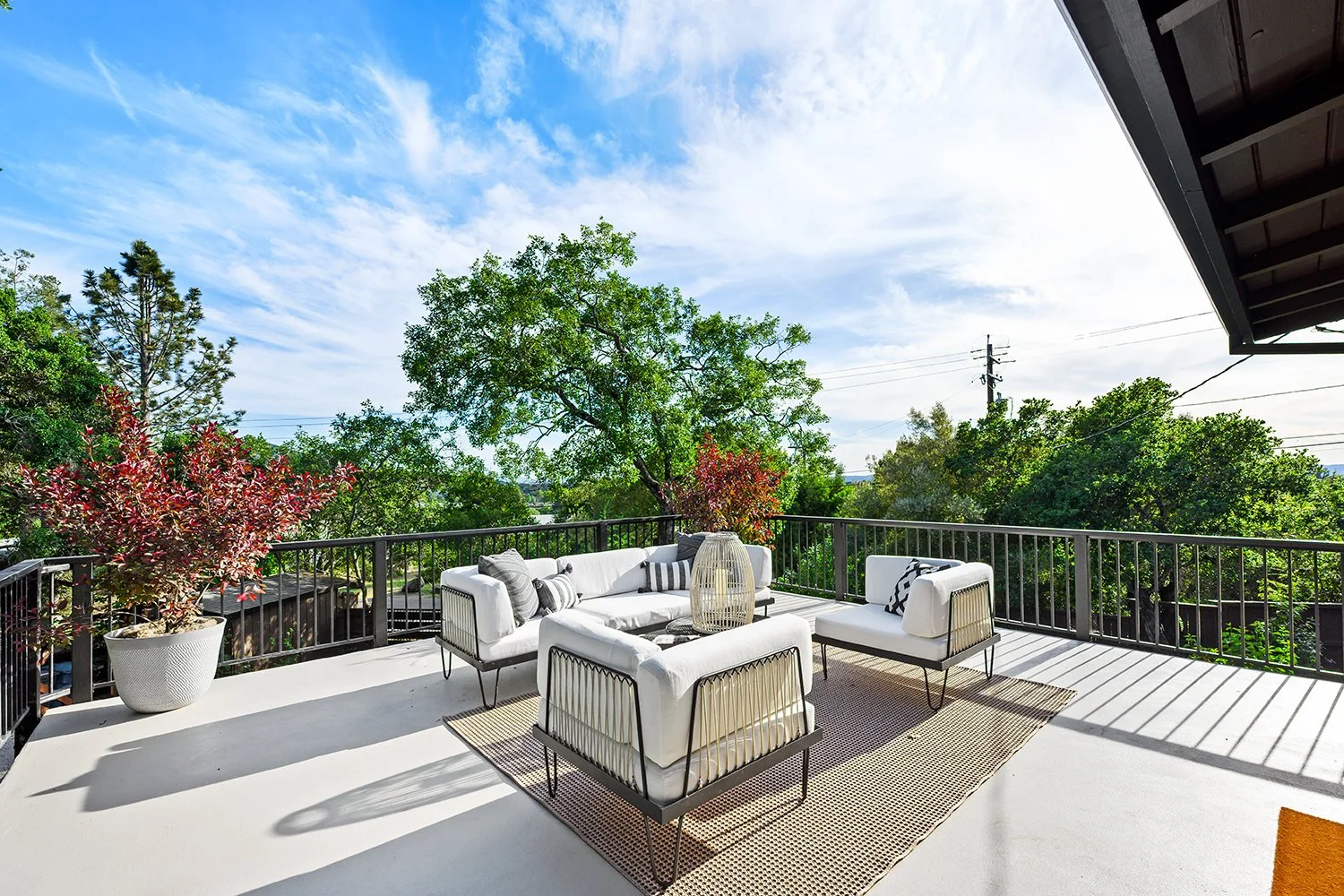 Outdoor balcony with white furniture, including a sofa and chairs, surrounded by potted plants and green trees under a partly cloudy sky.