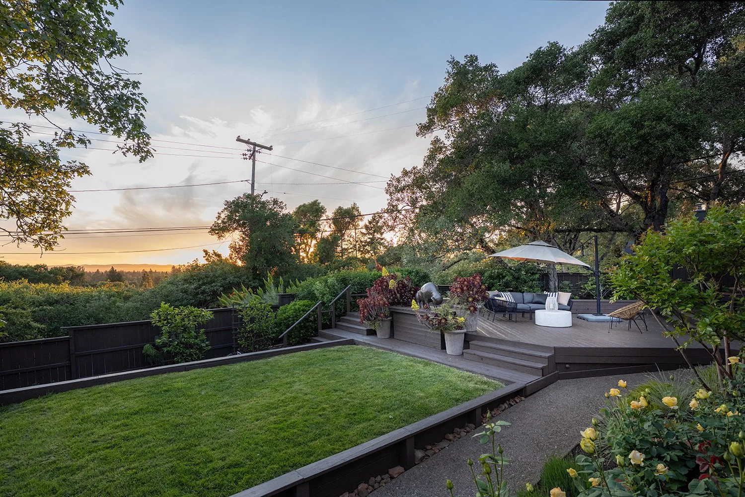 A backyard deck with outdoor furniture, including a sofa with an umbrella, lounge chairs, and potted plants, overlooking a lush green lawn and trees at sunset.
