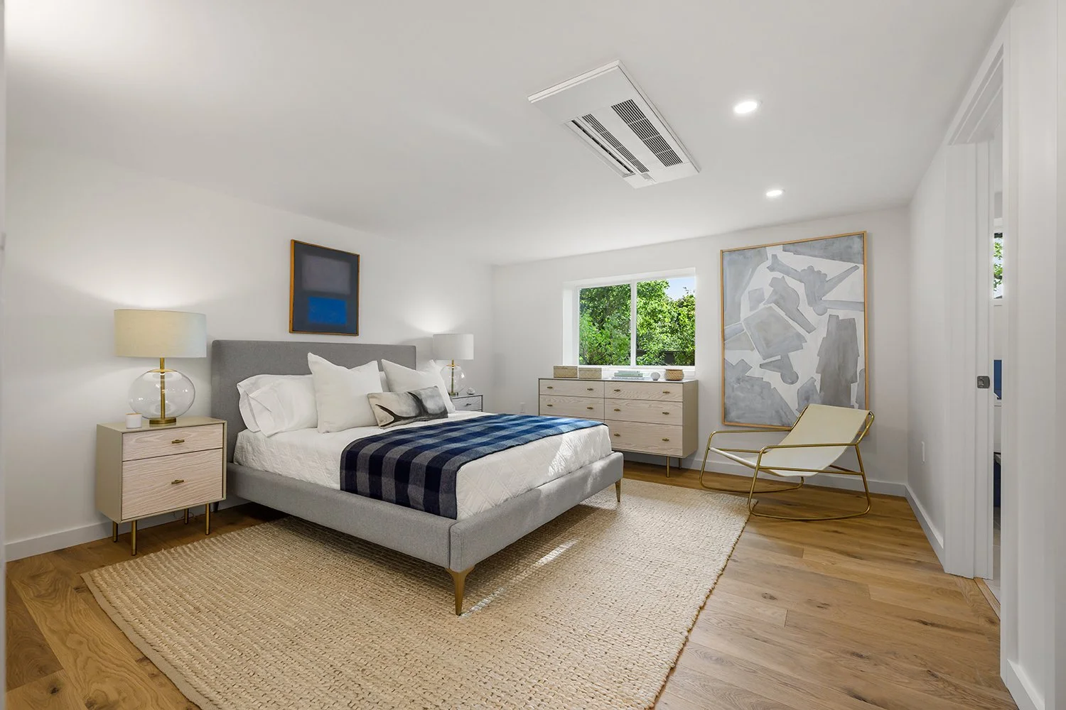 Modern bedroom with a gray bed, white bedding, and blue throw blanket. Features wooden nightstands with lamps, a white dresser, and abstract art. Wooden flooring with a large beige rug and a chair in the corner.