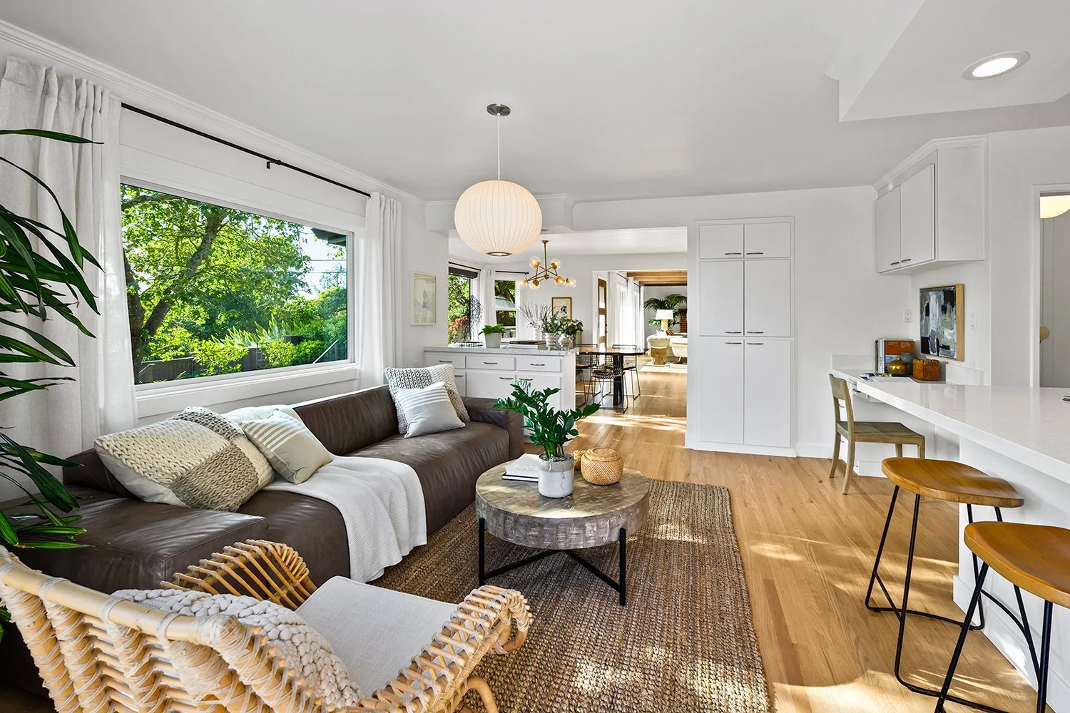 Bright living room with large window, brown leather sofa with beige and white pillows, rattan chair, round marble-top coffee table with a potted plant, hanging ceiling lights, white walls, hardwood floors, and a view into a dining area and kitchen.