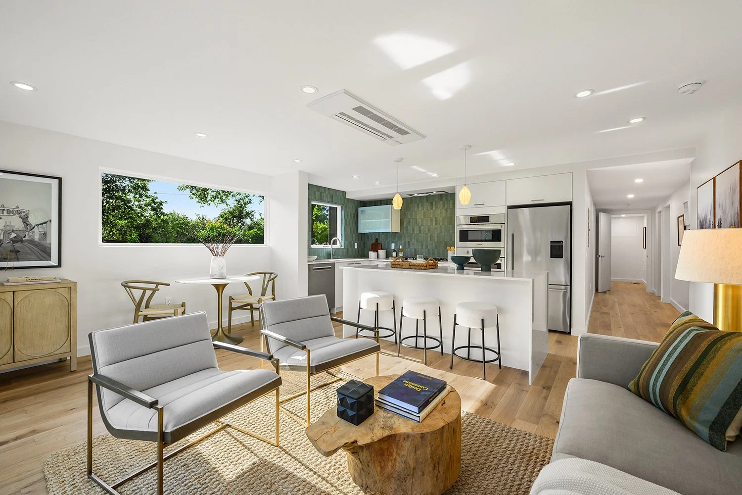Bright open-concept living room and kitchen with white walls, light wood flooring, modern furniture, large window with a view of greenery, and a kitchen with green textured backsplash and stainless steel appliances.