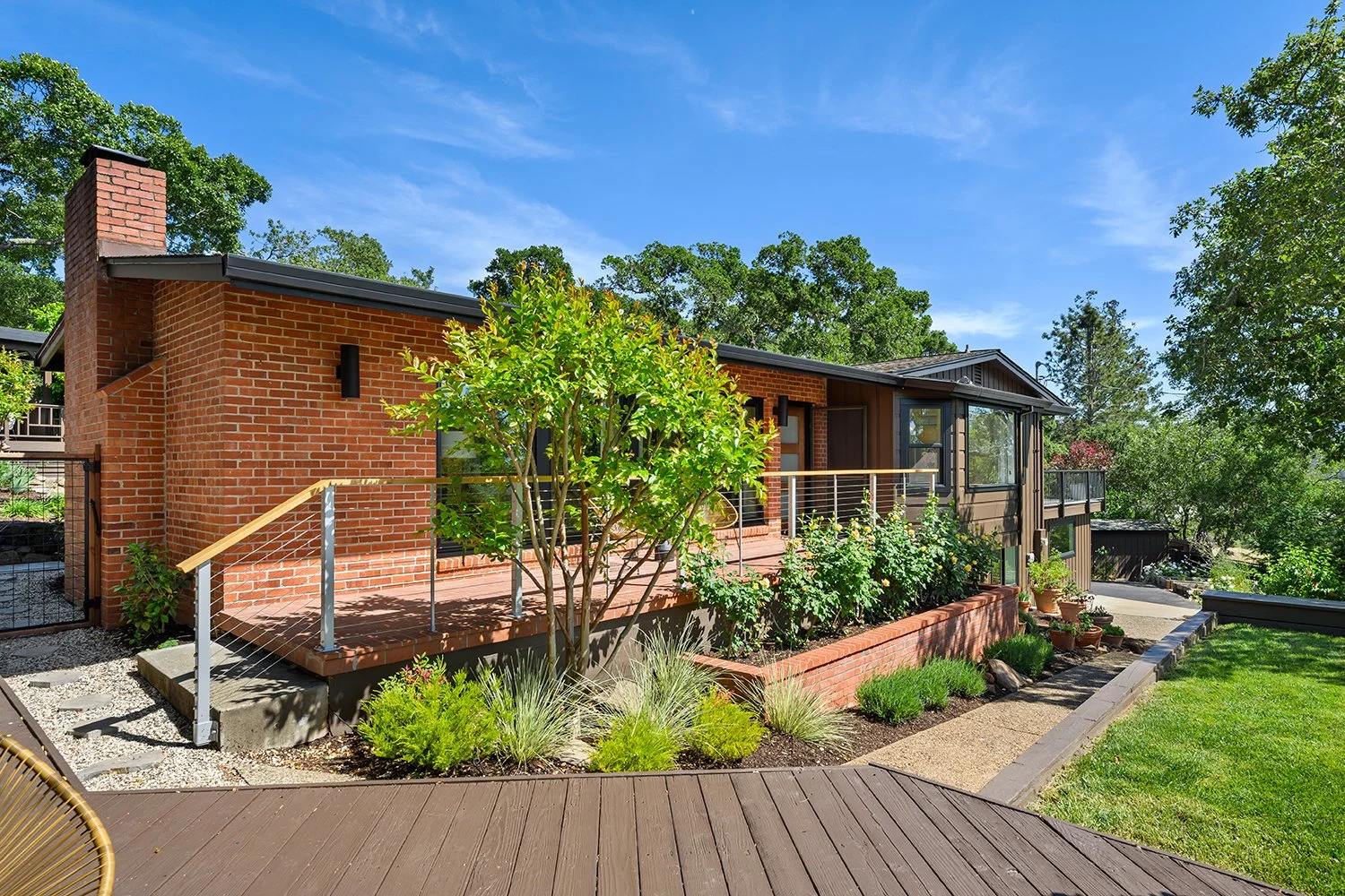 A brick house with a wooden deck and a garden with various plants and trees, under a clear blue sky.