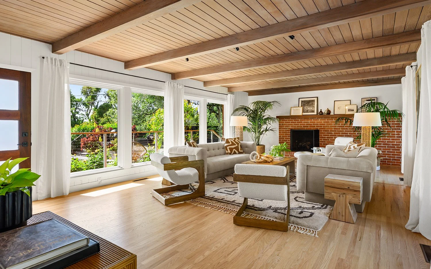 Bright living room with large windows showing garden, wood floor, white and beige furniture, brick fireplace, potted plants, and wooden ceiling beams.