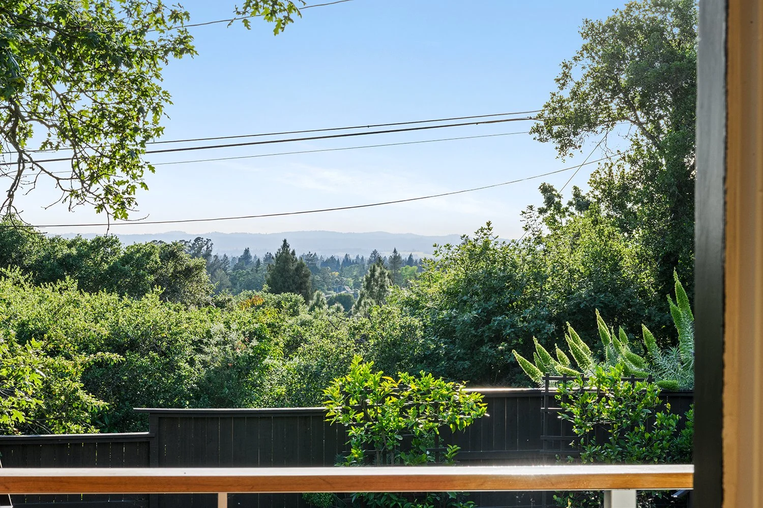 View of lush green trees and distant hills through a window with a wooden railing, under a clear blue sky.