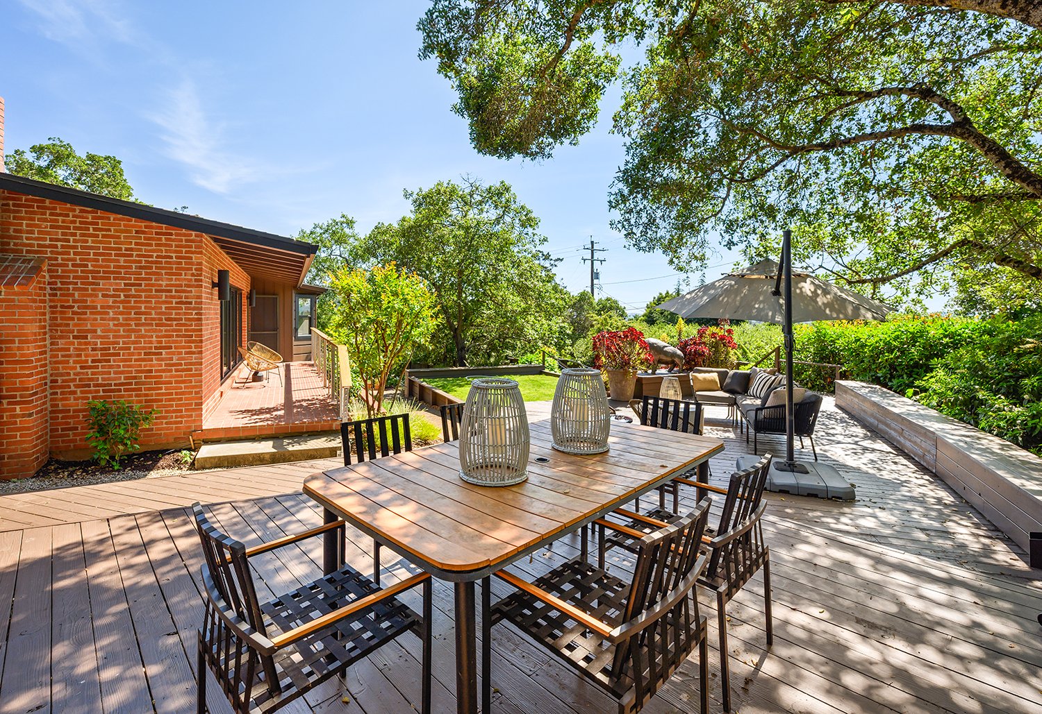 Outdoor patio with wooden deck, dining table with chairs, seating area with umbrella, plants, and trees in a backyard.