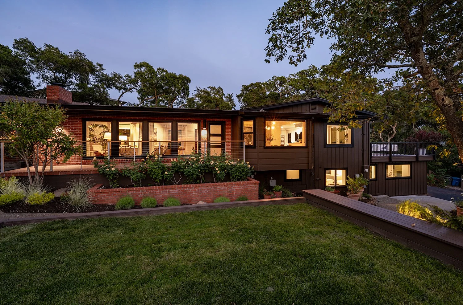 A modern house with a brick and dark wood exterior, illuminated interior lights, surrounded by a well-maintained garden and trees at dusk.
