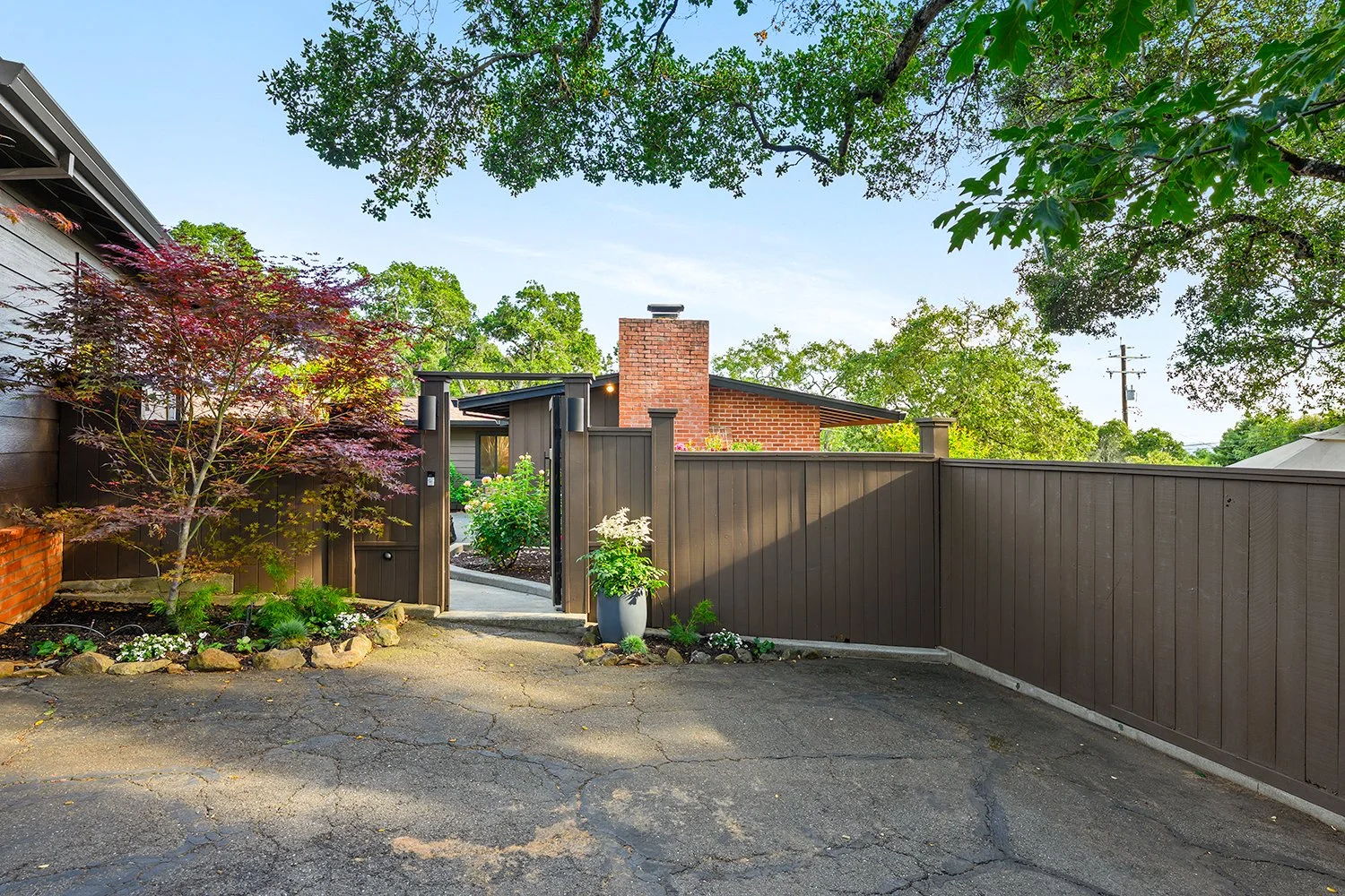 A backyard with a tall, brown wooden fence, an open gate, a flower bed with trees and plants, and a partially visible house with a brick chimney. Trees and a power line are visible in the background.