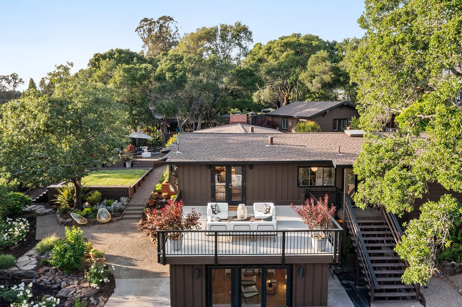 A backyard view of a modern house with a balcony, outdoor furniture, trees, and landscaped garden.