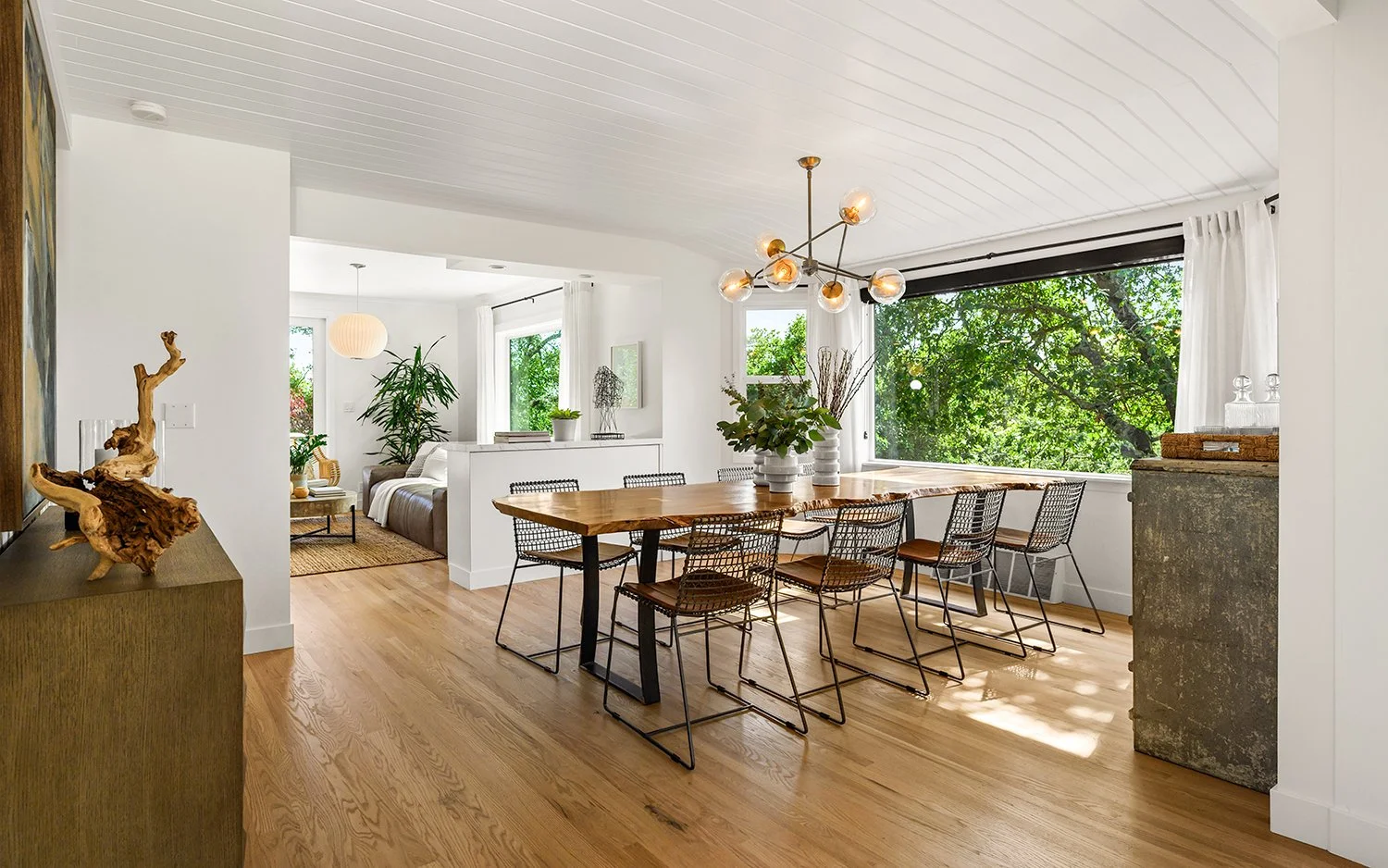 Bright dining room with large window, wooden table, eight black wire chairs, pendant light fixture, and a view of green trees outside.
