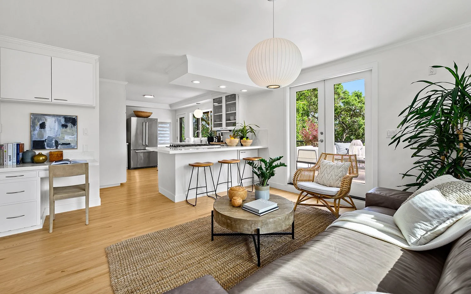 Bright living room with white walls, wooden flooring, a sofa with pillows, round coffee table with books and decorative items, a wicker chair with a cushion, large window and glass door showing outdoor patio and trees, kitchen with white cabinets, a kitchen island with bar stools, stainless steel refrigerator, and hanging paper lantern light.