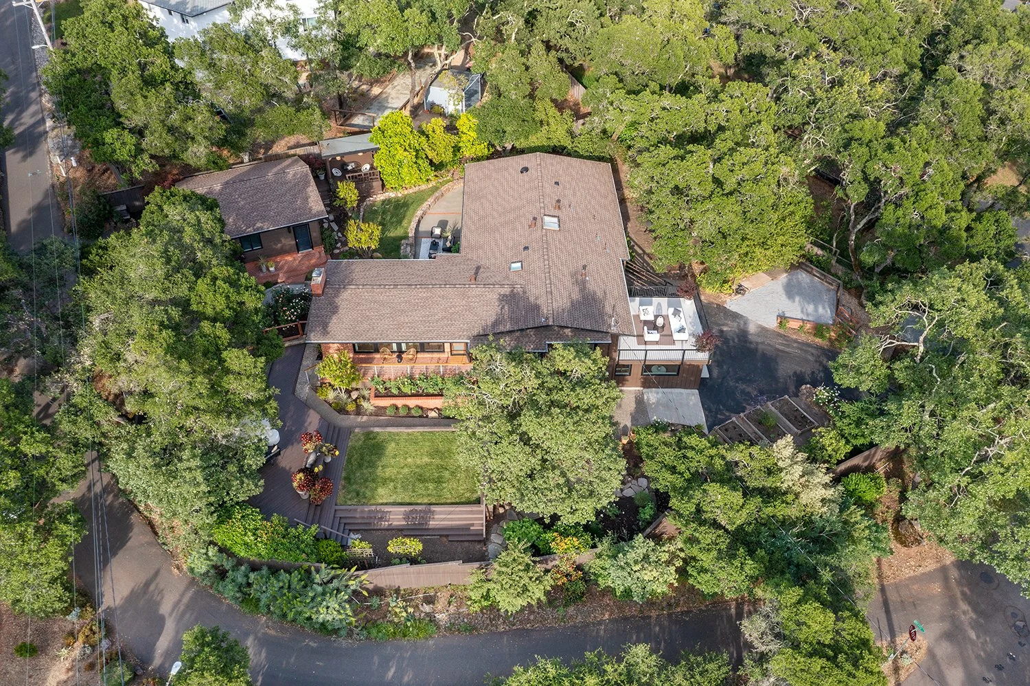 An aerial view of a house surrounded by trees and greenery, with a paved driveway, a deck with outdoor furniture, and a small lawn area.