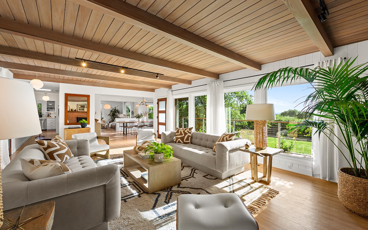 Bright living room with large windows, white sofas with patterned pillows, a beige rug, wooden coffee table with potted plant, wooden ceiling with exposed beams, and a view of the greenery outside.