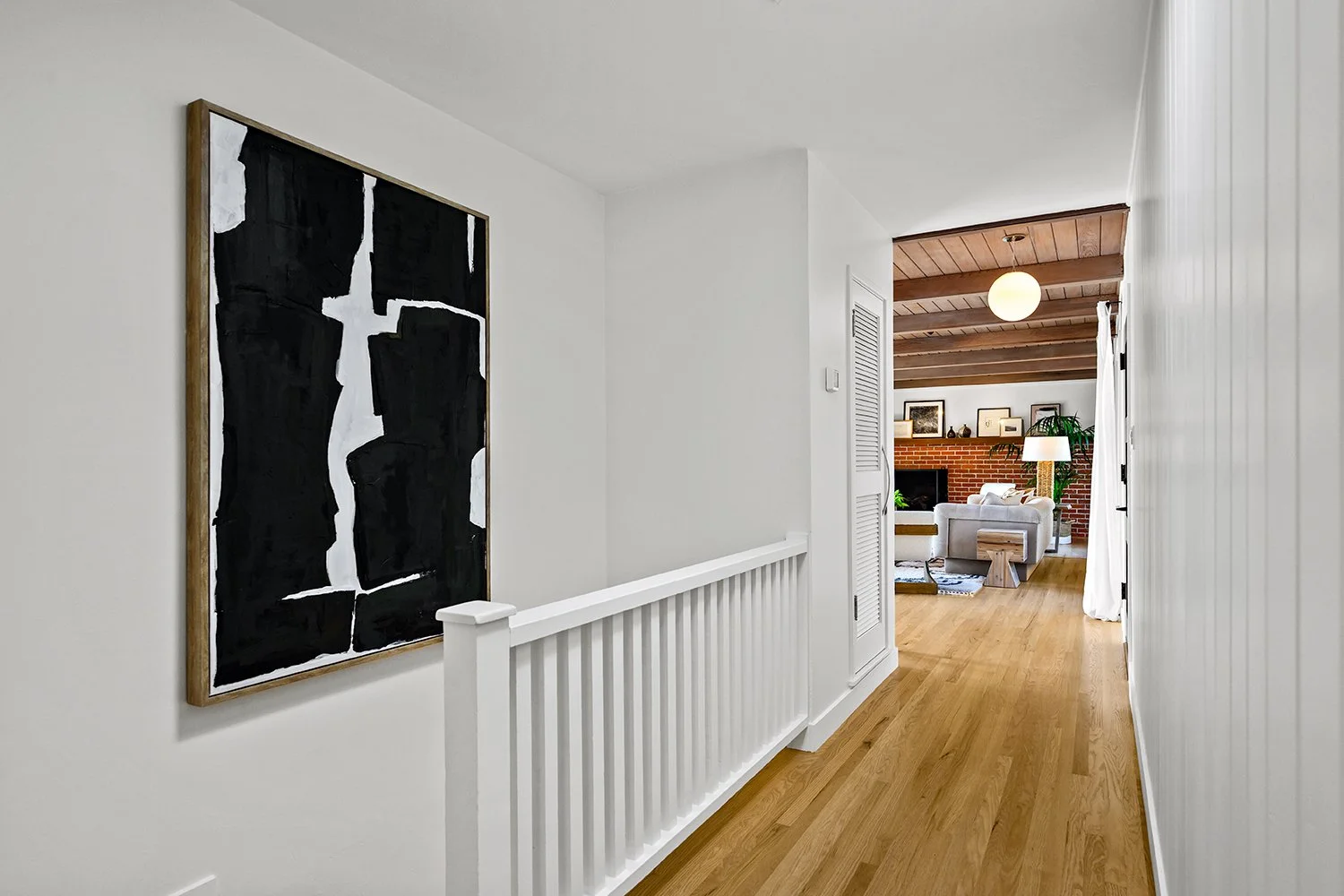 Hallway with white walls, featuring a framed abstract black and white painting on the left, leading to a living room with a brick wall, wooden ceiling, white sofa, and decorative items.