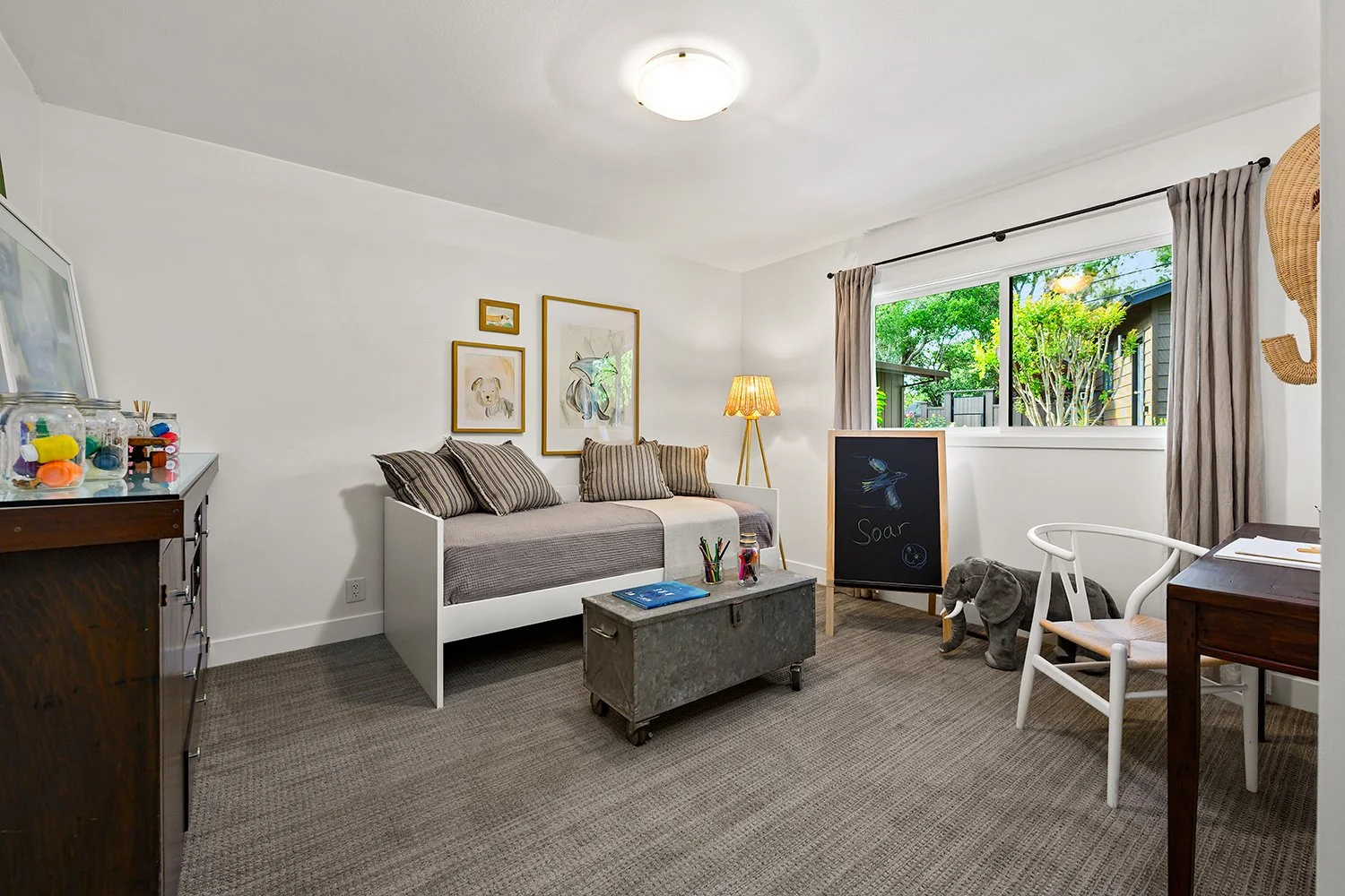 Bright, cozy living room with white walls, a large window with beige curtains, and a gray carpet. Features include a white daybed with striped pillows, a small table with art supplies, a chalkboard with a bird drawing and the word 'Soar,' a house-sha