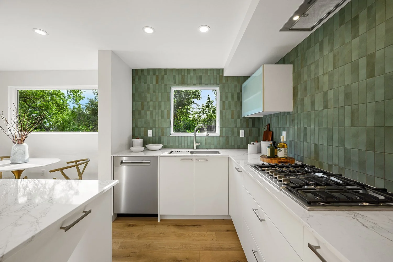 Modern kitchen with white cabinets, a green tiled backsplash, a window showing trees outside, and a gas stove.