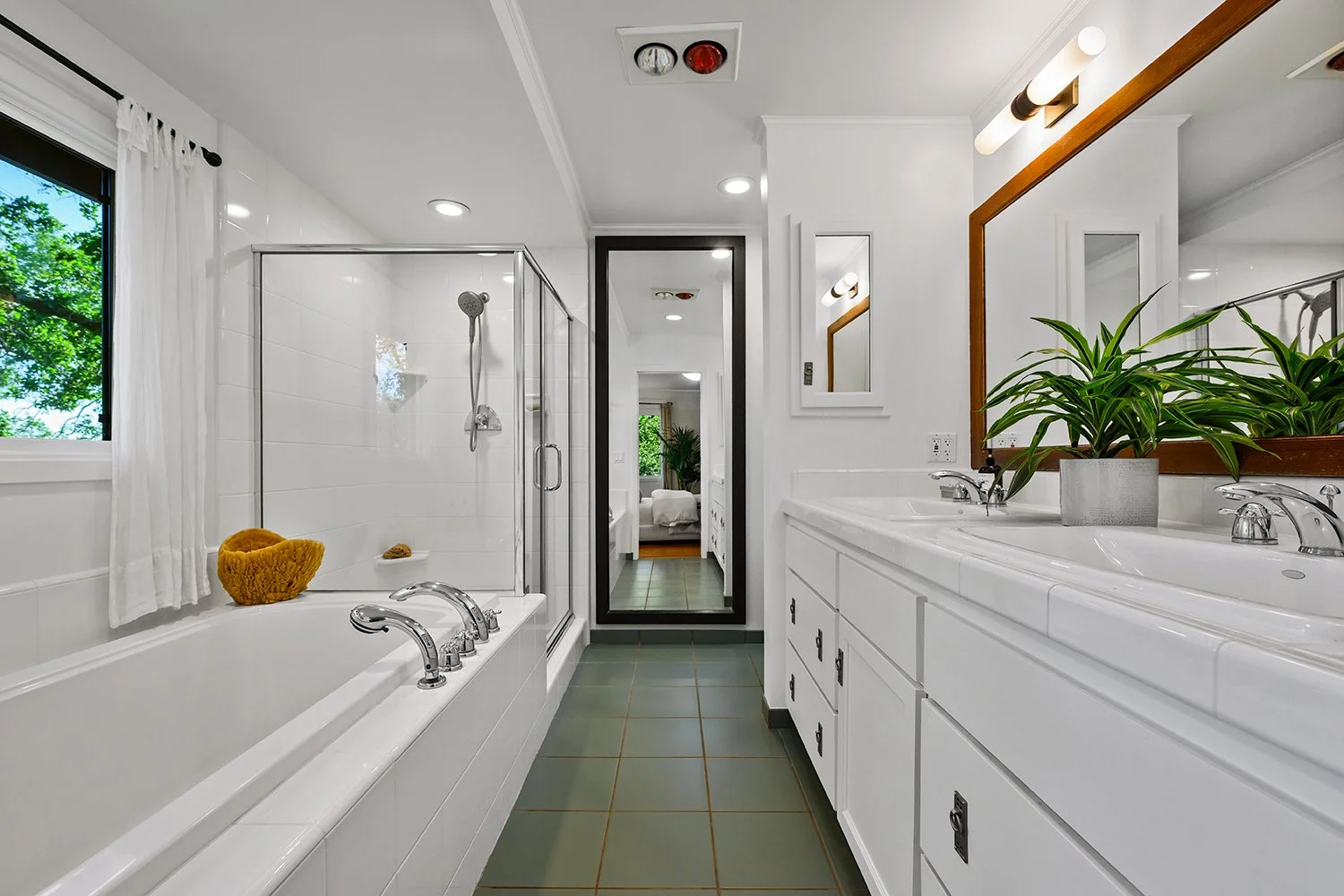 White bathroom with window, bathtub, glass shower stall, double sink vanity, mirror, potted plant, and bedroom visible through open door.