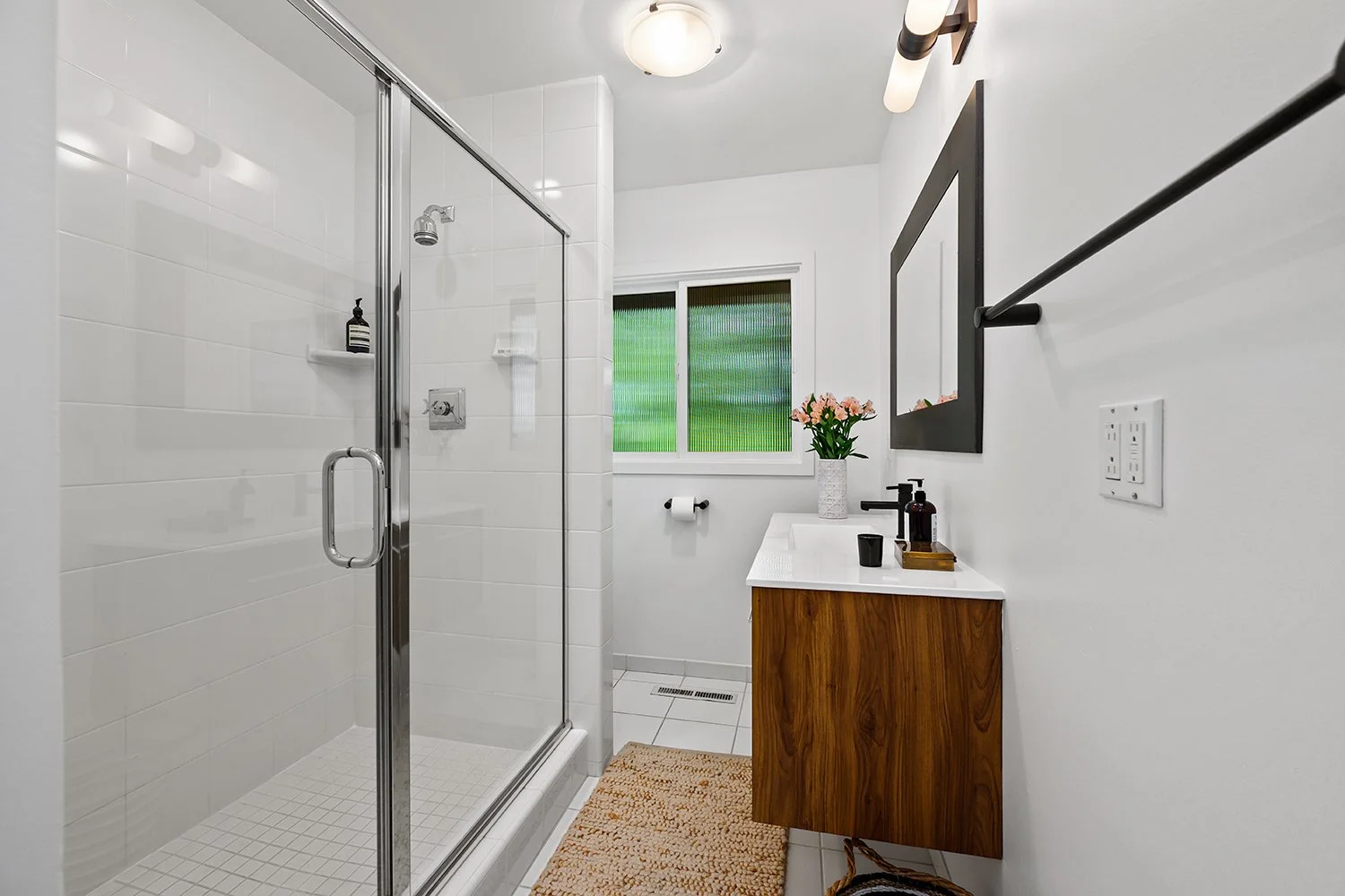 A modern bathroom with a glass shower enclosure, a white wall, a wooden vanity with a white countertop, a black faucet, a mirror with black frame, a window with textured glass, a potted plant with pink flowers, a towel bar, a black cup, soap dispense