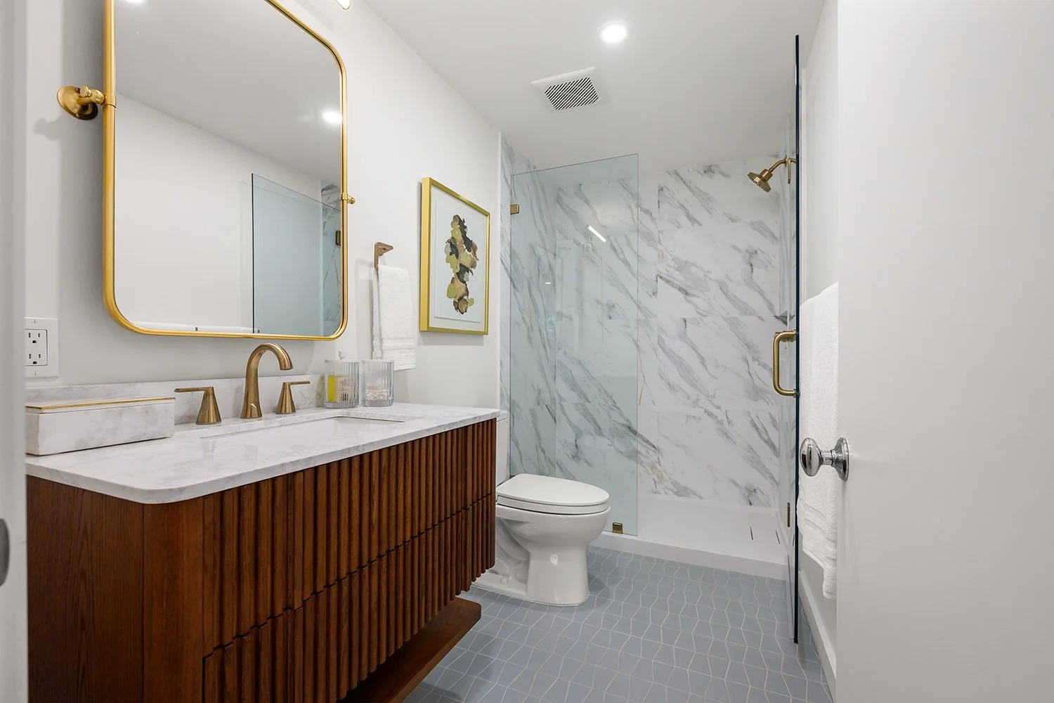 Modern bathroom with marble shower, wooden vanity with marble countertop, gold fixtures, mirrored wall, and framed artwork.