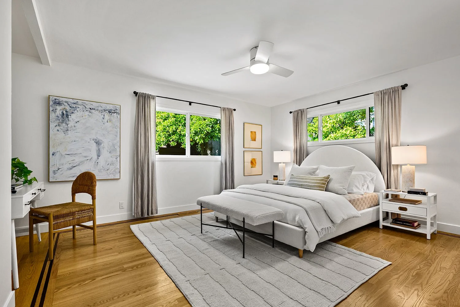 A bright bedroom with white walls, a bed with white linens and pillows, two bedside tables with lamps, an abstract painting on the wall, a wicker chair in the corner, and windows with beige curtains showing green trees outside.