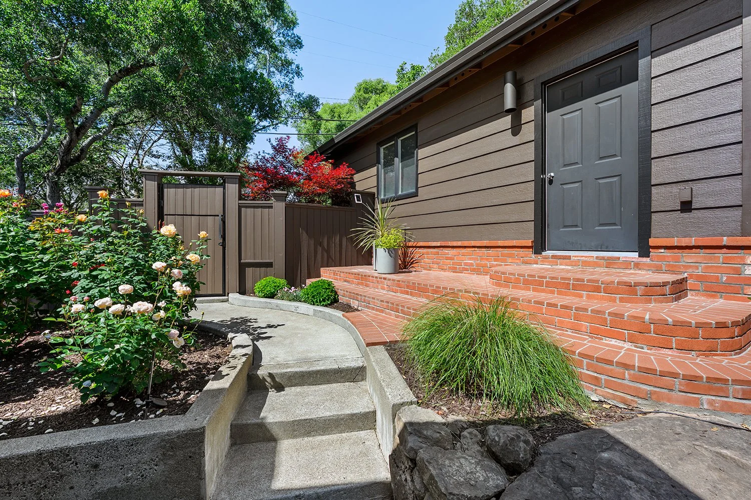 Front entrance of a house with brick steps, brown siding, black door, and landscaped garden with roses and green plants.
