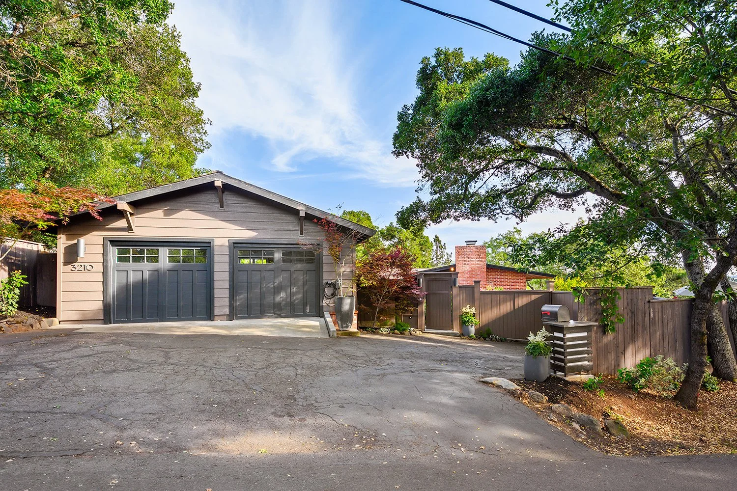 A driveway leading to a gray garage with two doors, surrounded by green trees and plants, with a wooden fence and mailbox on the right side.