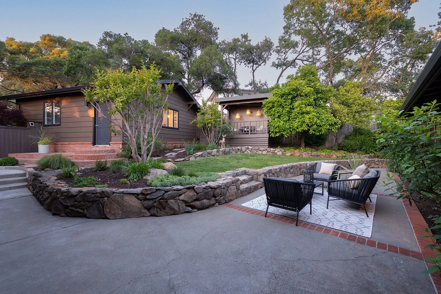 A backyard patio area with outdoor seating, a grassy landscaped area, and a small house or shed in the background with trees and bushes surrounding the space.