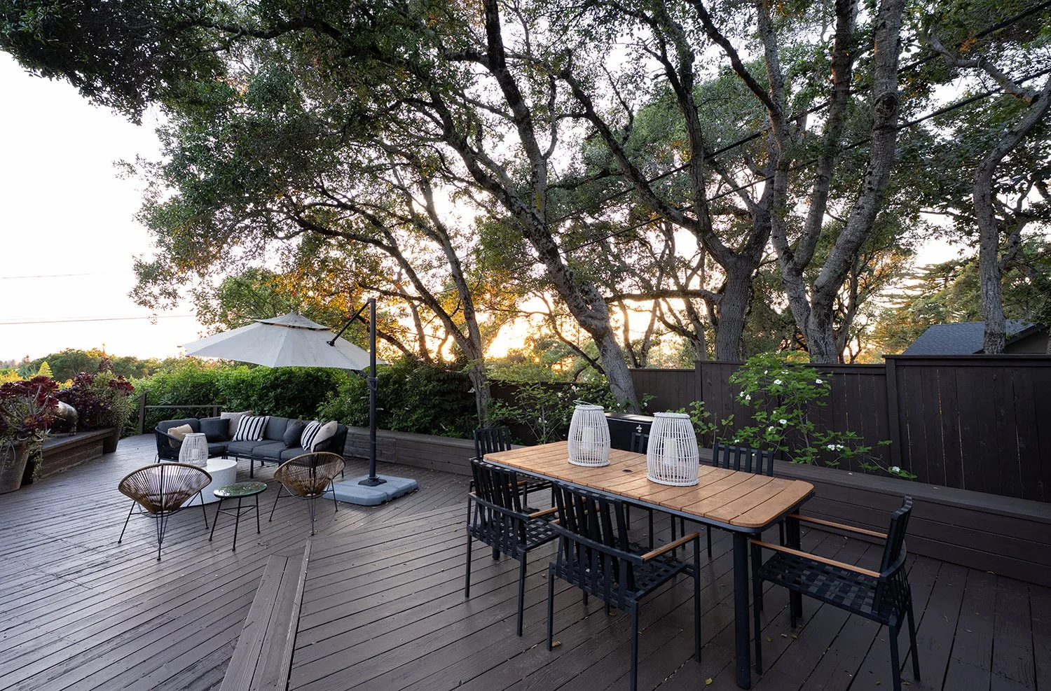 Outdoor wooden patio with black chairs, a wooden table with decorative lanterns, a black cushioned outdoor sofa with striped pillows, a large white umbrella, and lush green trees in the background. Sunset light.