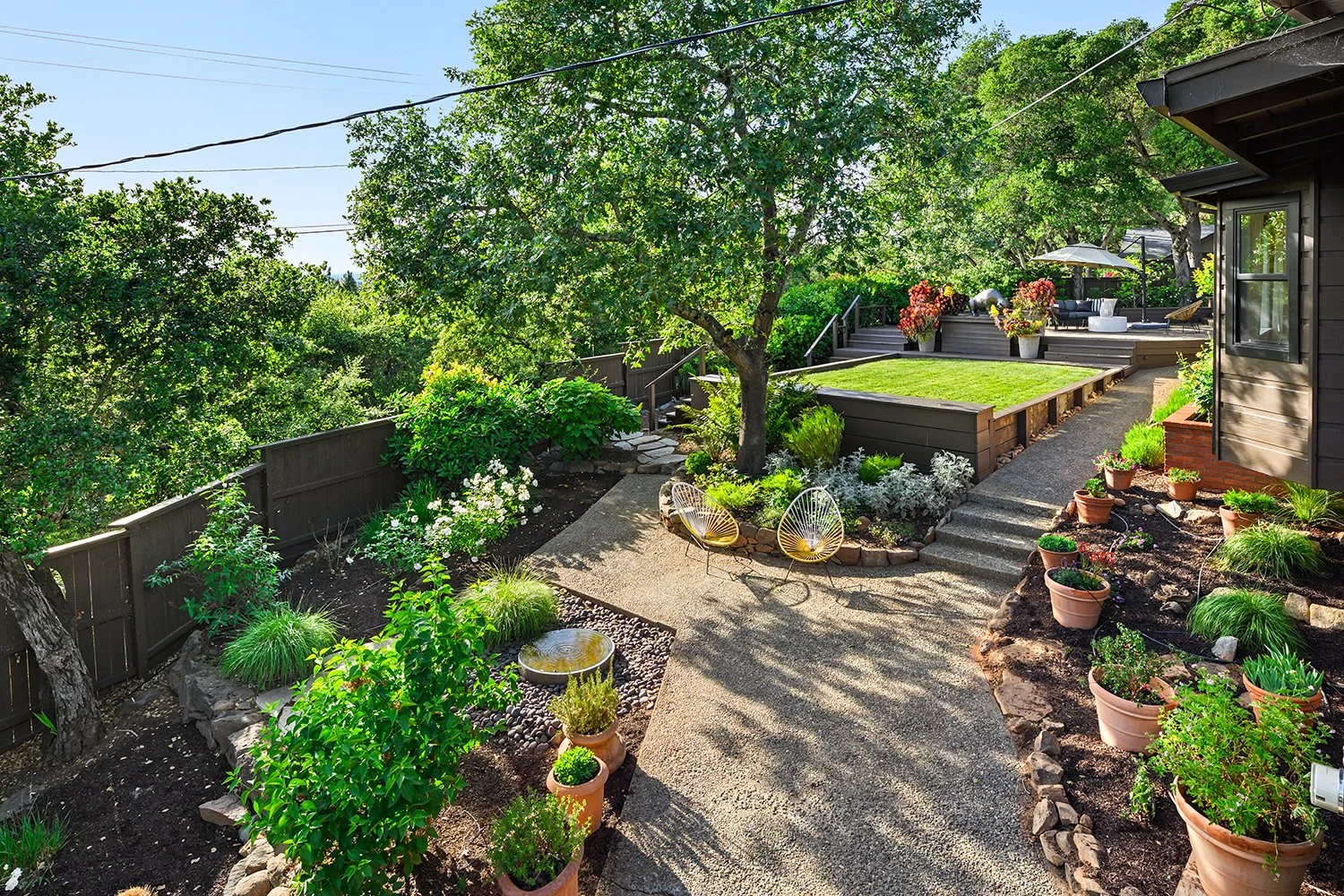 A lush, well-maintained backyard garden with potted plants, a stone pathway, a large tree, a raised grass area, outdoor seating with a patio umbrella, and blooming flowers.