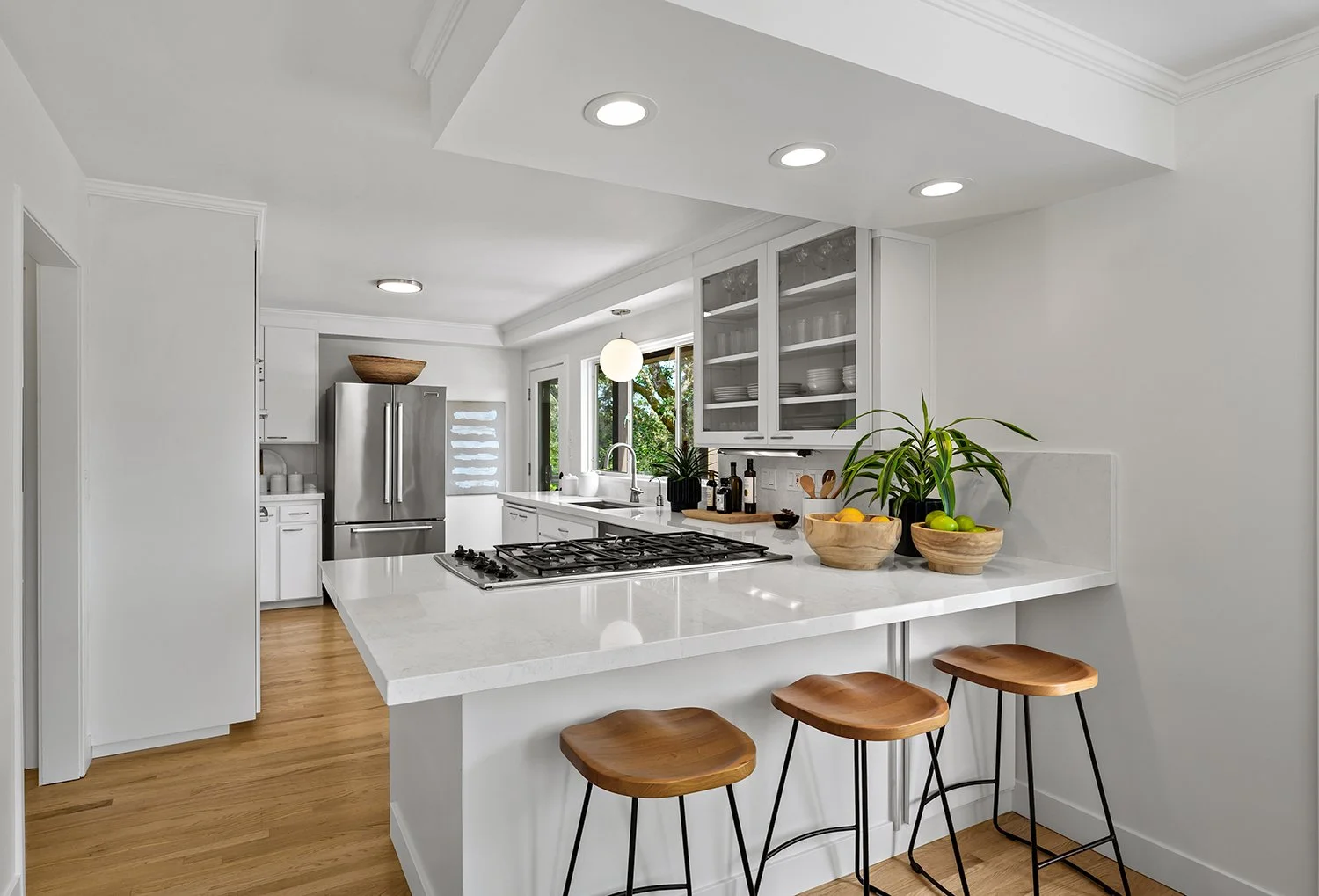 Modern white kitchen with wooden bar stools, stainless steel refrigerator, white countertops, glass-front cabinets, and natural light from windows.