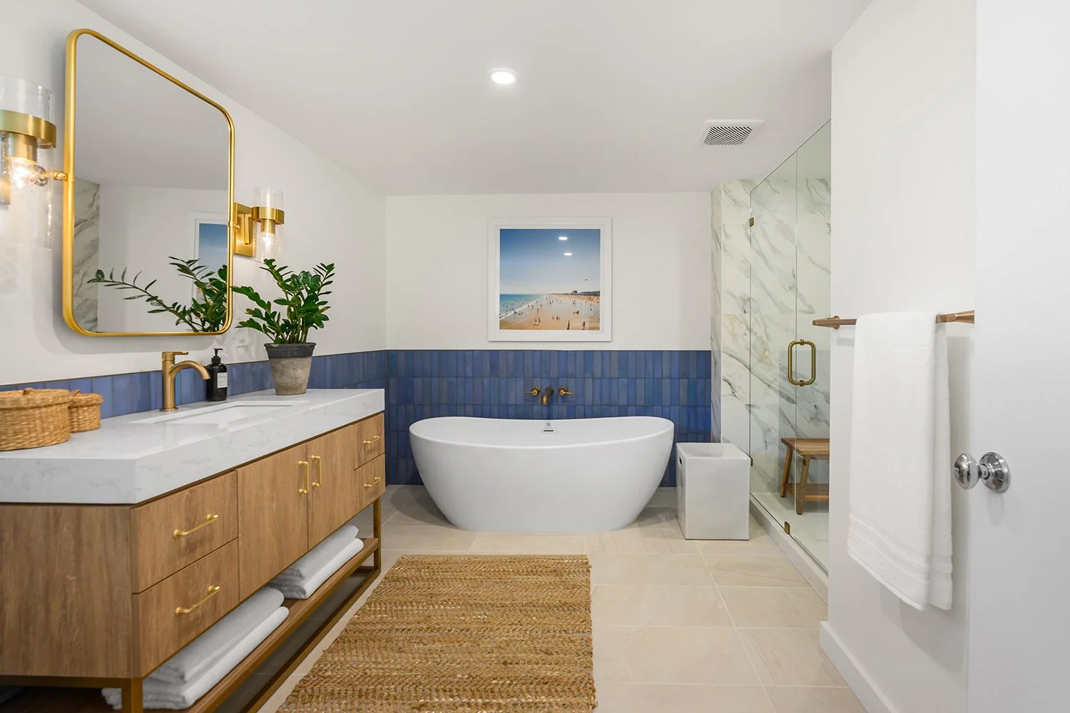 Modern bathroom with a freestanding bathtub, blue tiled wall, wooden vanity with marble countertop, large mirror with gold frame, potted plant, framed beach photo, glass shower with marble walls, white towels, and beige tile flooring.