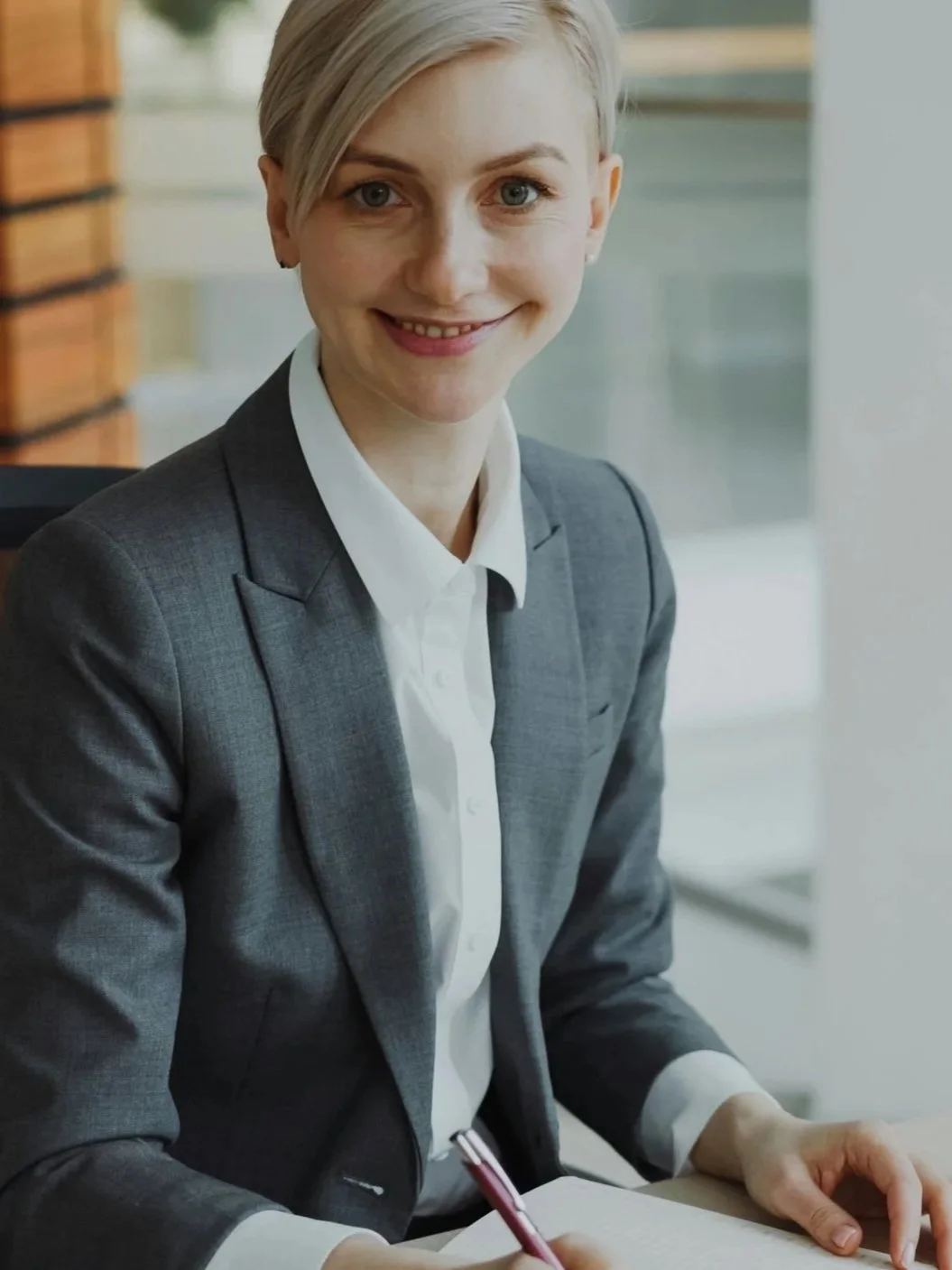 A professional woman with short blonde hair, wearing a gray blazer and white shirt, sitting at a desk and smiling at the camera in an office setting.