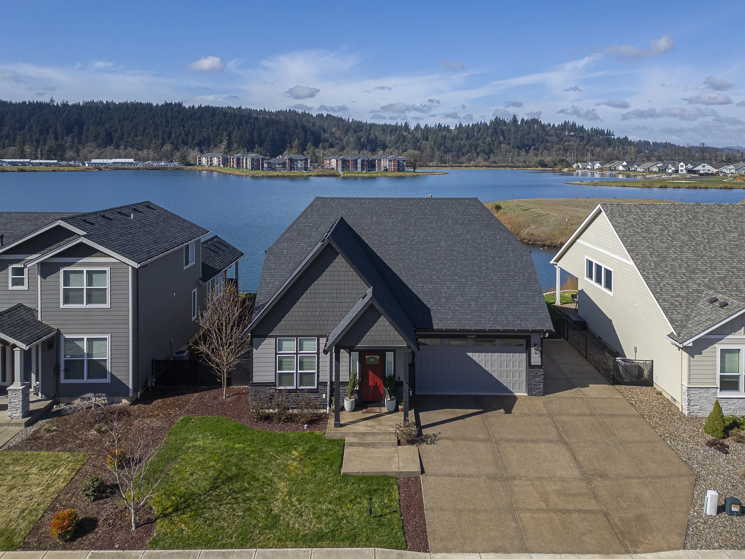 Aerial view of a craftsman-style home with red front door and two-car garage backing to a lake with forested hills in Salem, Oregon