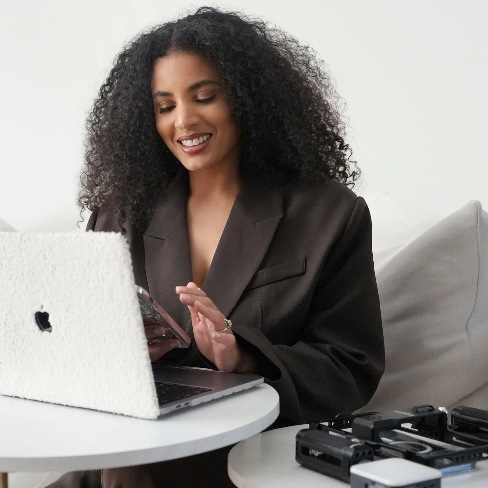 A woman with long, curly black hair, smiling while looking at her phone, sitting at a white table with a laptop in front of her.