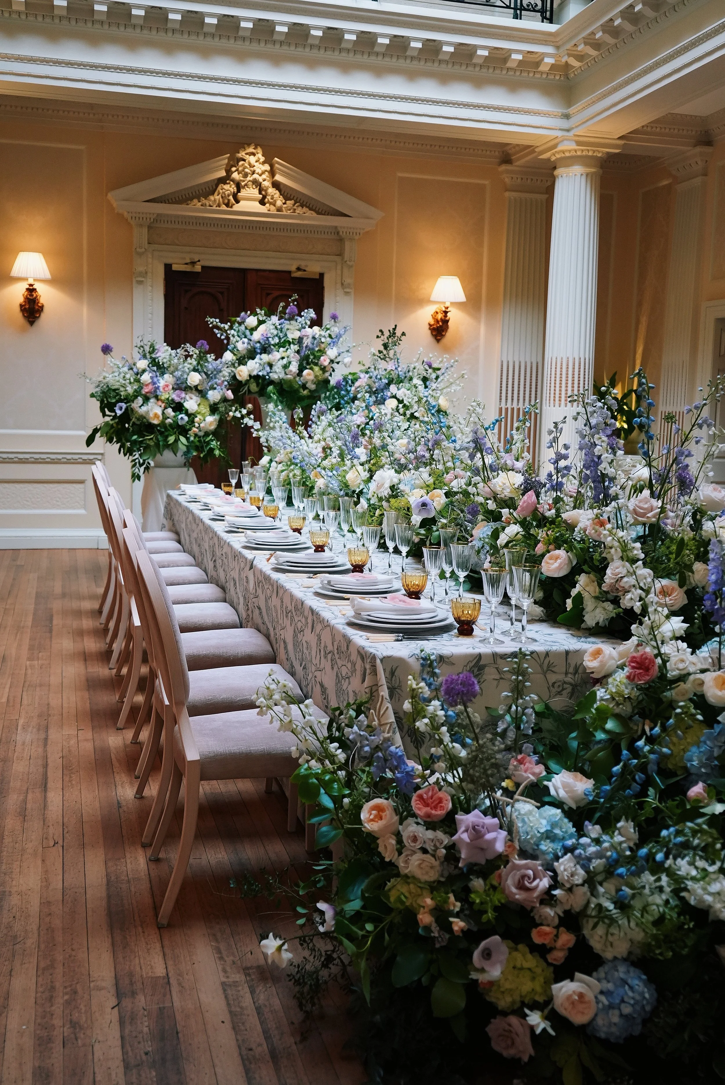 Elegant banquet table decorated with a large abundance of pastel-colored flowers in shades of pink, purple, white, and blue, set in a grand room with ornate architectural details, wall sconces, and soft lighting.