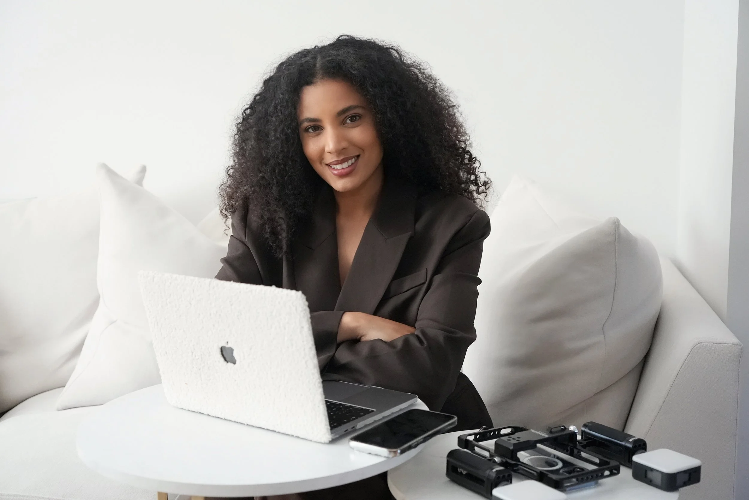 A woman with curly black hair smiling while working on a laptop in a white room with white pillows.