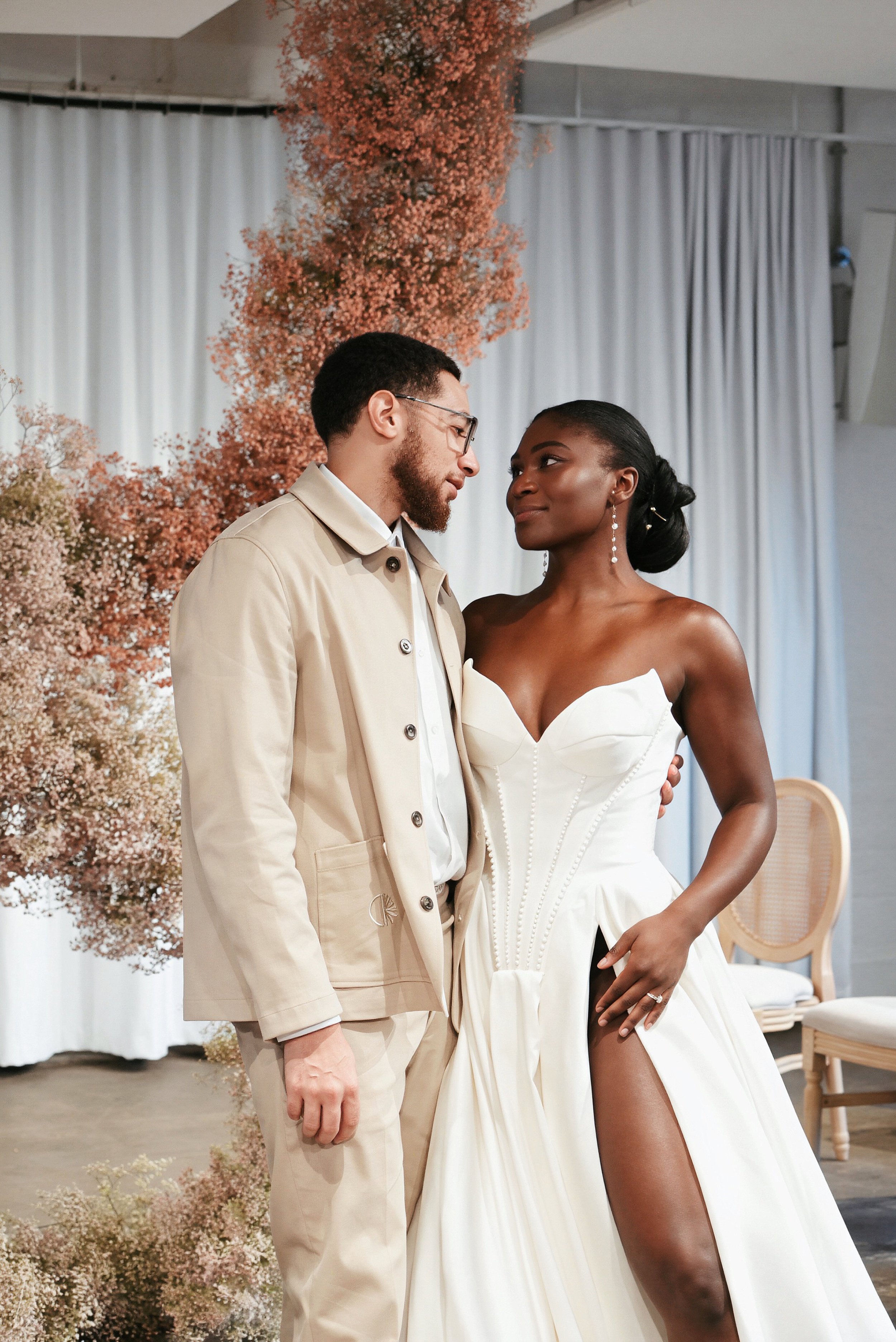A couple getting married in a wedding ceremony, standing close and looking into each other's eyes. The man is wearing glasses and a beige trench coat, while the woman is in a white wedding dress with a high slit, showing her leg. There are pink flowers and white curtains in the background.