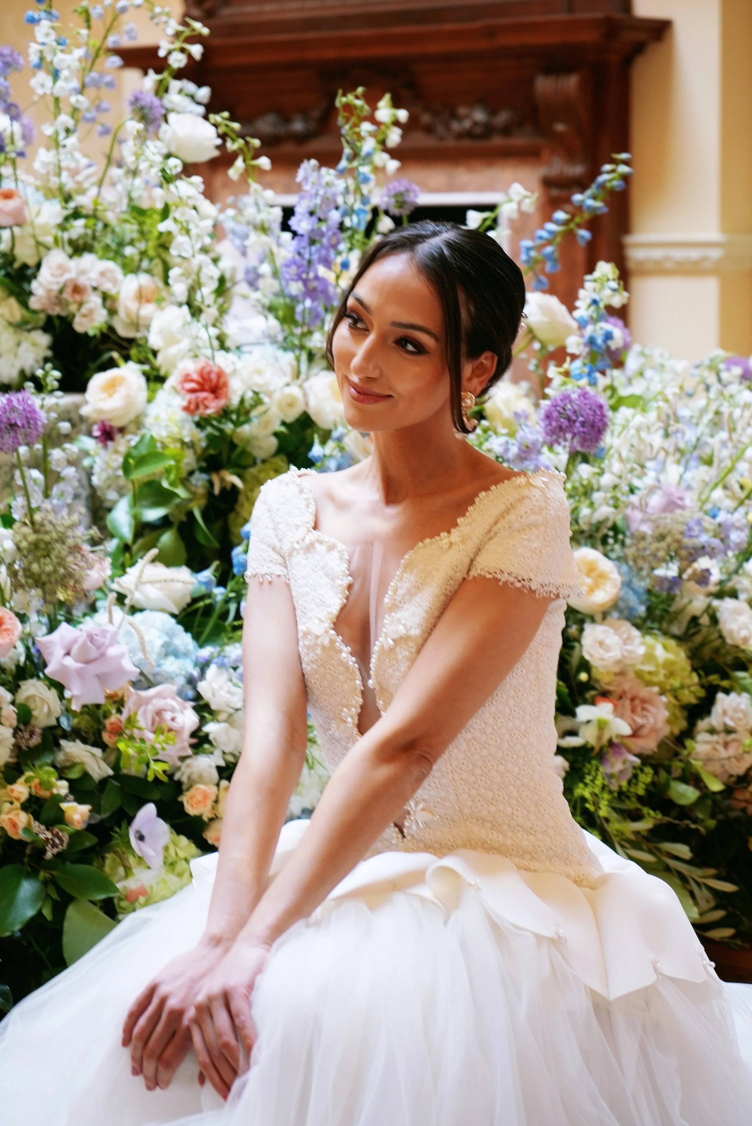 A woman in a wedding dress seated in front of a large floral arrangement with various flowers in pastel and white colors.